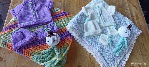 Two sets of hand-knitted baby clothes and blankets are displayed on a wooden table, each including cardigans, hats, booties, and crochet comfort dolls, arranged on colorful and pastel-patterned blankets.