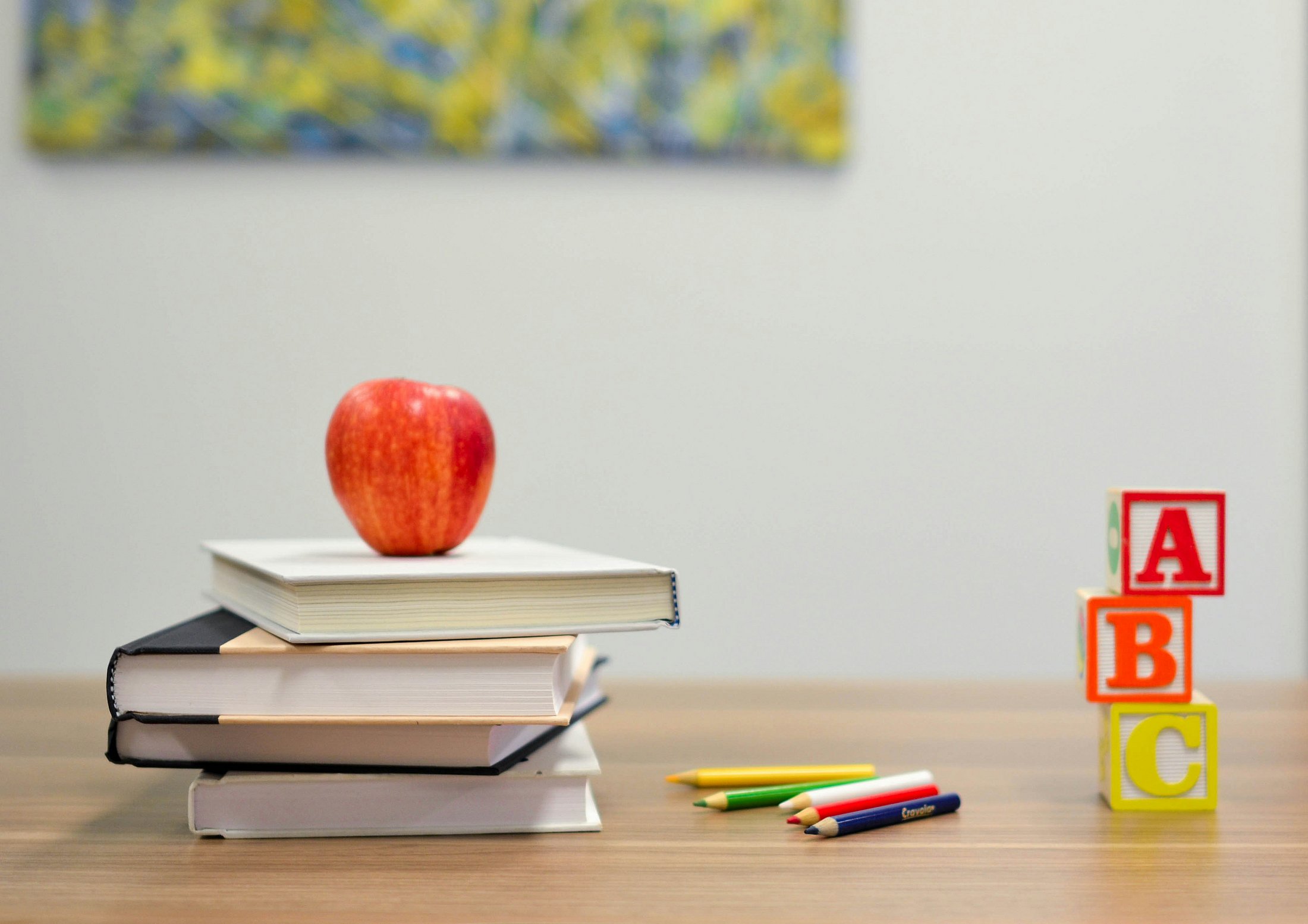A stack of books with a red apple on top sits beside alphabet blocks and colored pencils on a table. The image represents educational outcomes and lifelong learning in the context of preterm birth and child development.
