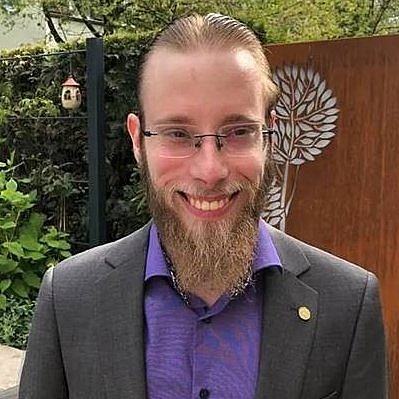 A professional headshot of Lukas Mader, smiling in a suit with a garden background.