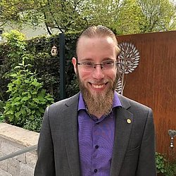 A professional headshot of Lukas Mader, smiling in a suit with a garden background.