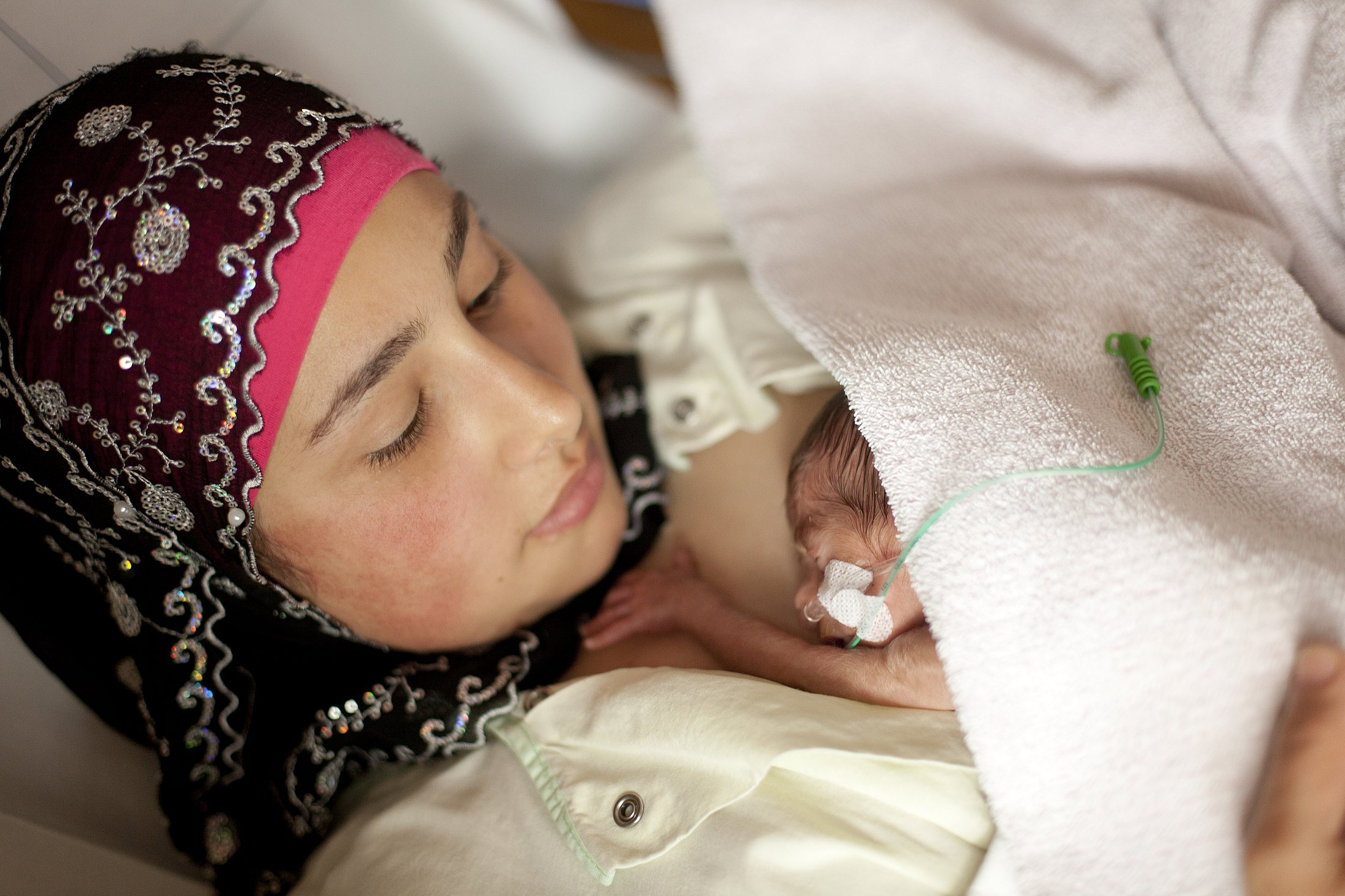 Mother and Preterm Infant During Skin-to-Skin Care Mother wearing a decorated headscarf holds her preterm infant against her chest beneath a blanket.