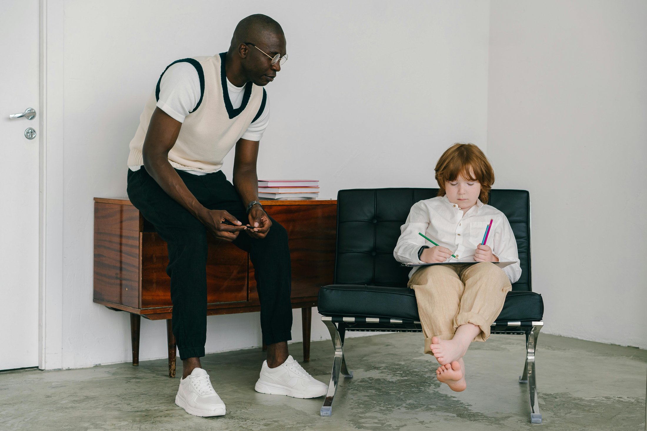 A man leans forward from a wooden cabinet, engaging with a barefoot child seated on a black chair who is drawing with colored pencils in a minimalist indoor setting.