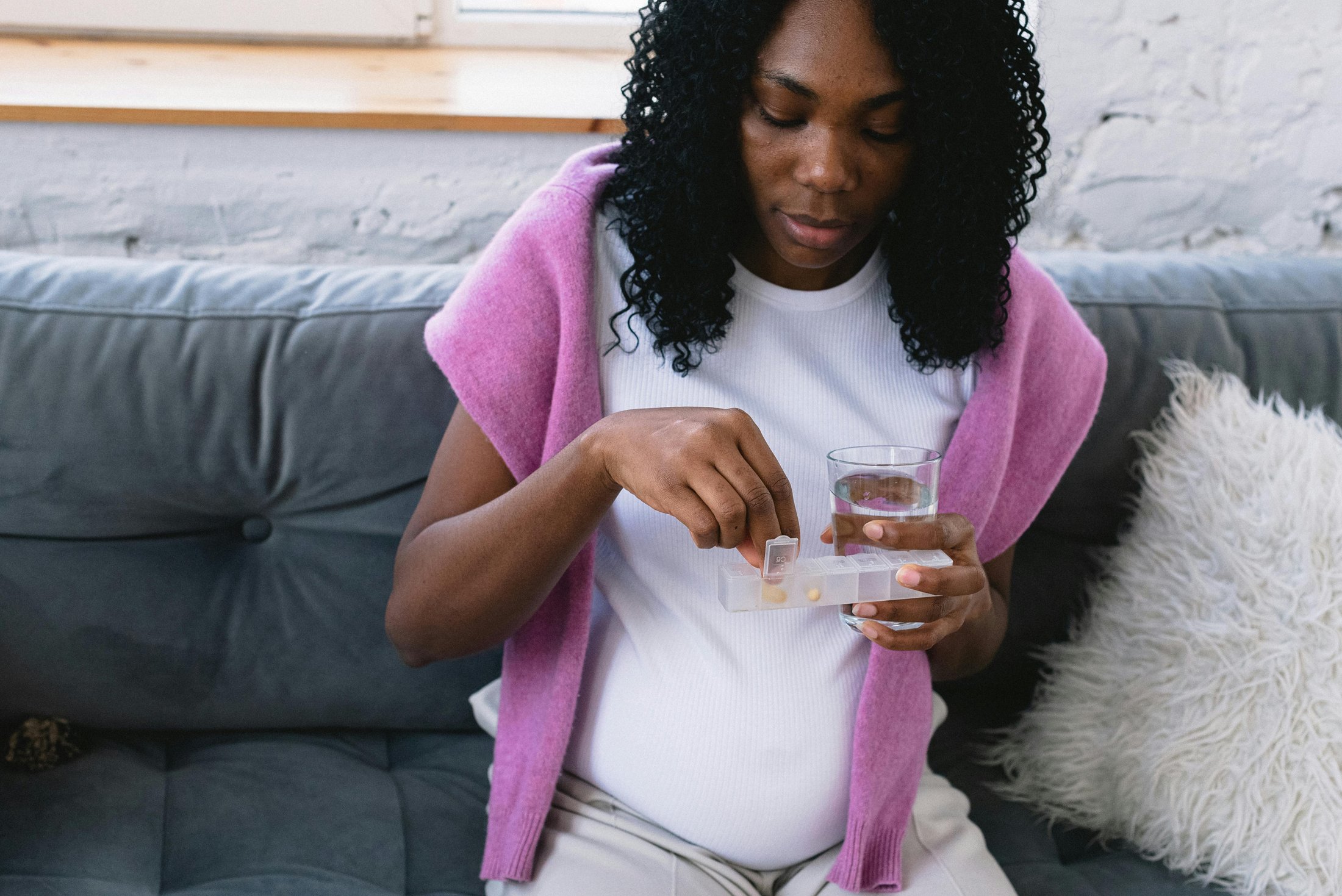 A pregnant woman sits on a couch holding a glass of water and a pill organizer. She is selecting a pill, possibly prenatal vitamins or prescribed medication, wearing a white shirt and a pink sweater draped over her shoulders.