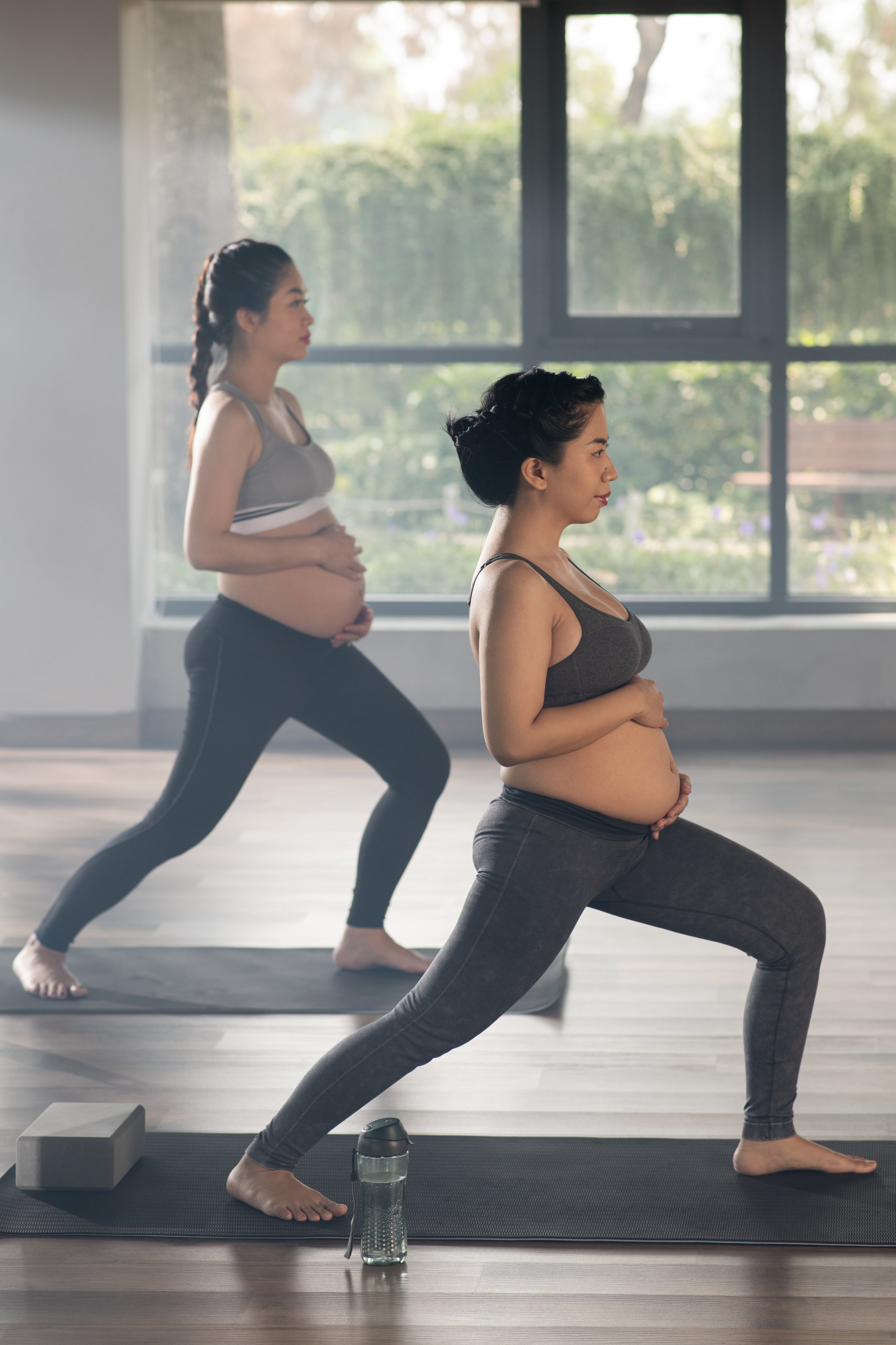 The image shows two pregnant individuals performing a low-impact exercise in an indoor studio with natural light, reflecting safe and guided physical activity during pregnancy and supportive care environments.