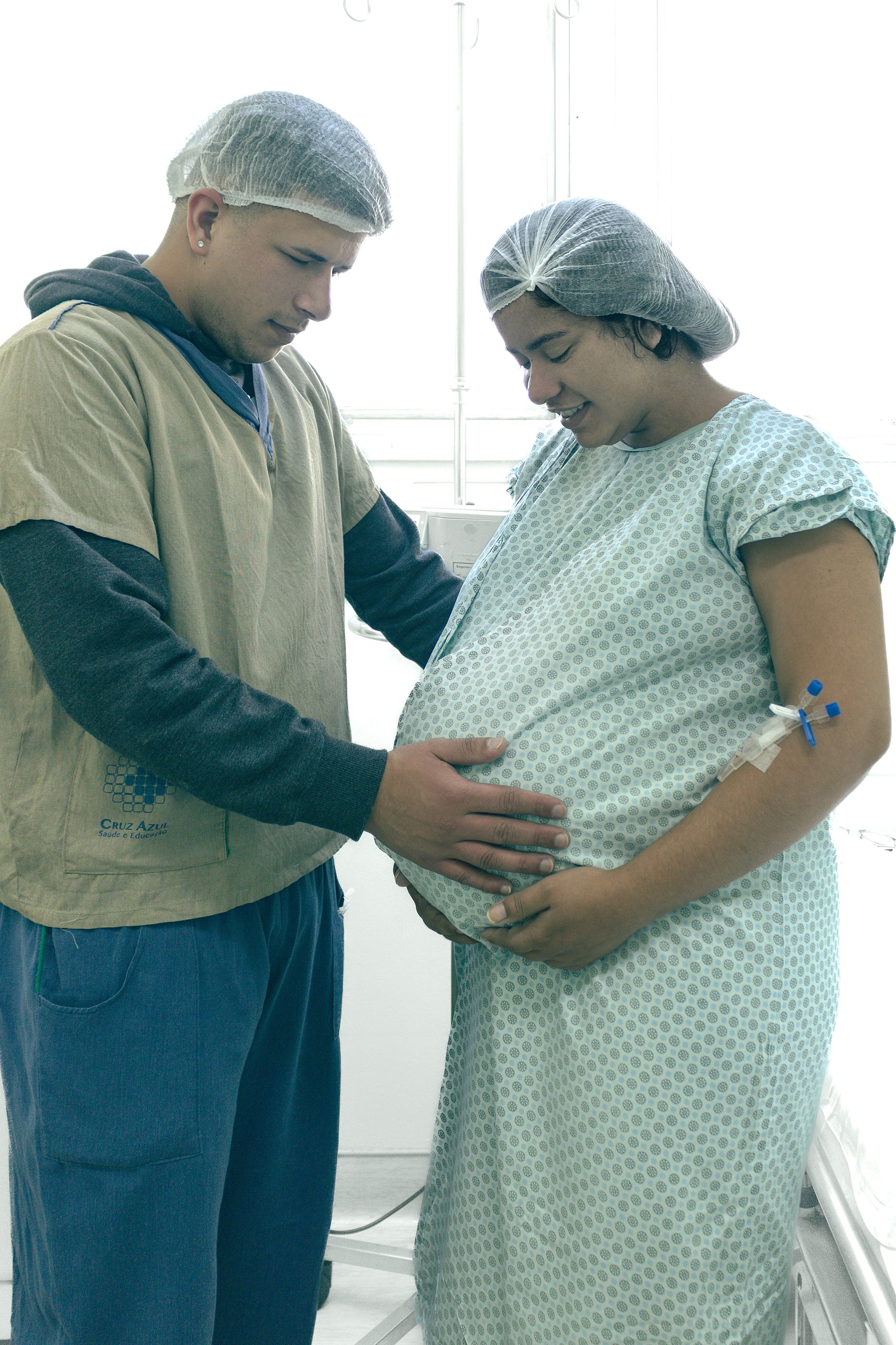 Partner Supporting a Pregnant Woman in Hospital Pregnant woman in a hospital gown standing with her partner, who rests a hand on her belly.