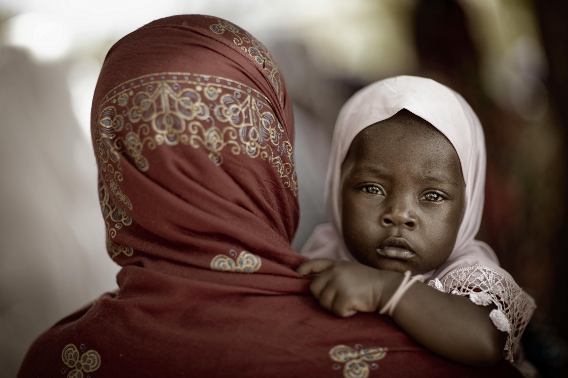 A close-up photograph of a young child resting against the shoulder of a woman wearing a red patterned headscarf. The child looks directly into the camera with a calm, thoughtful expression, while the woman is seen from behind.