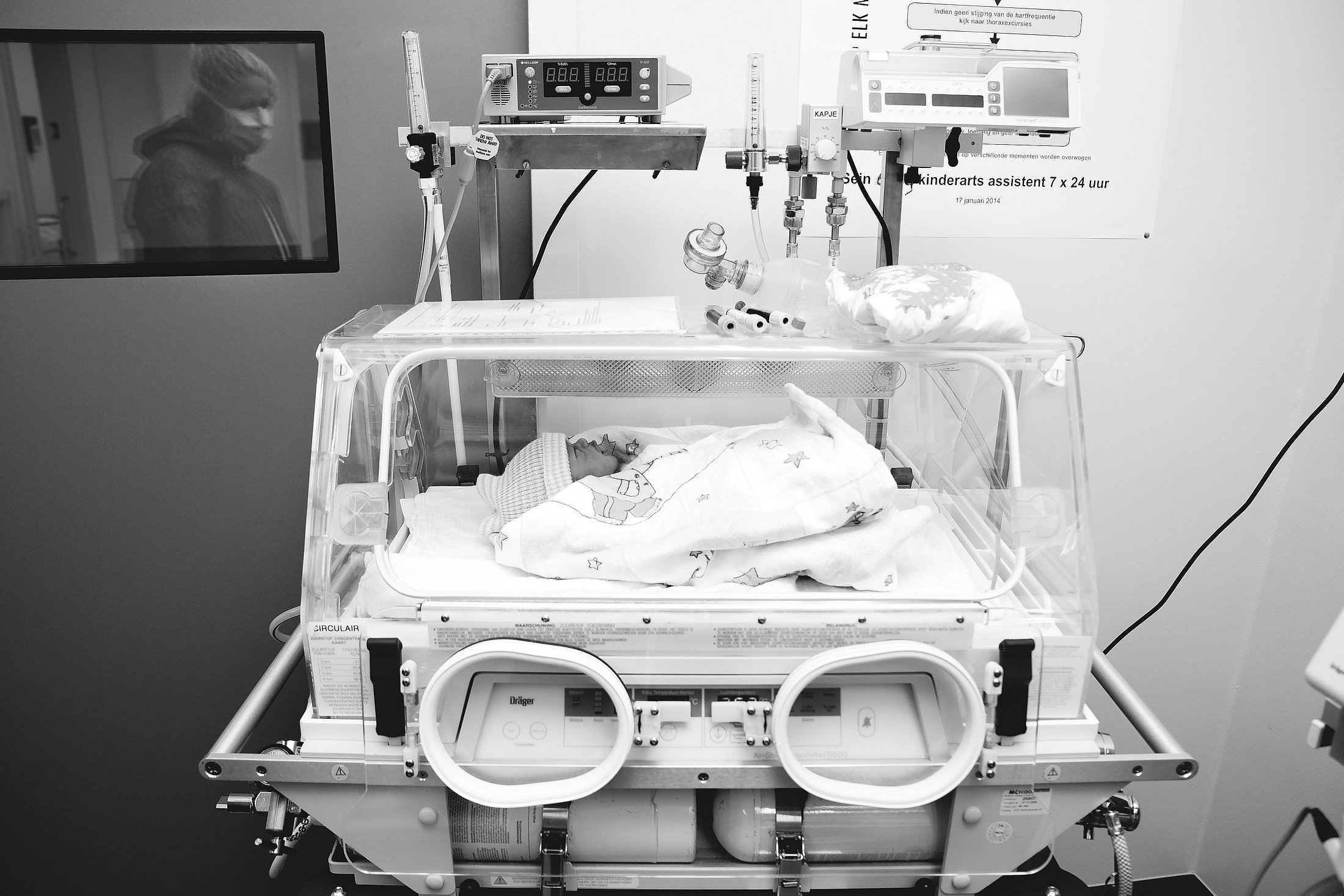 Neonatal Intensive Care and Nurse Workload A newborn infant rests in an incubator in a neonatal intensive care unit while a family member is visible through a window in the background. The image illustrates neonatal intensive care and highlights the importance of appropriate nurse workload to support safe, reliable care.