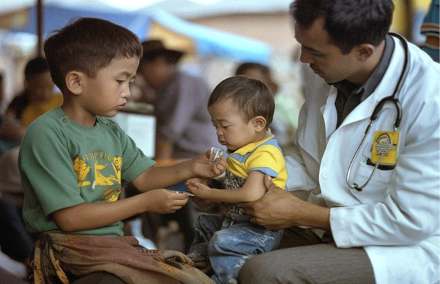 A doctor in a white coat assisting a young child in drinking medicine while an older child helps.