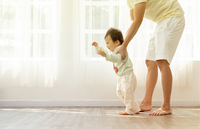 A toddler taking their first steps with parental support in a bright room.