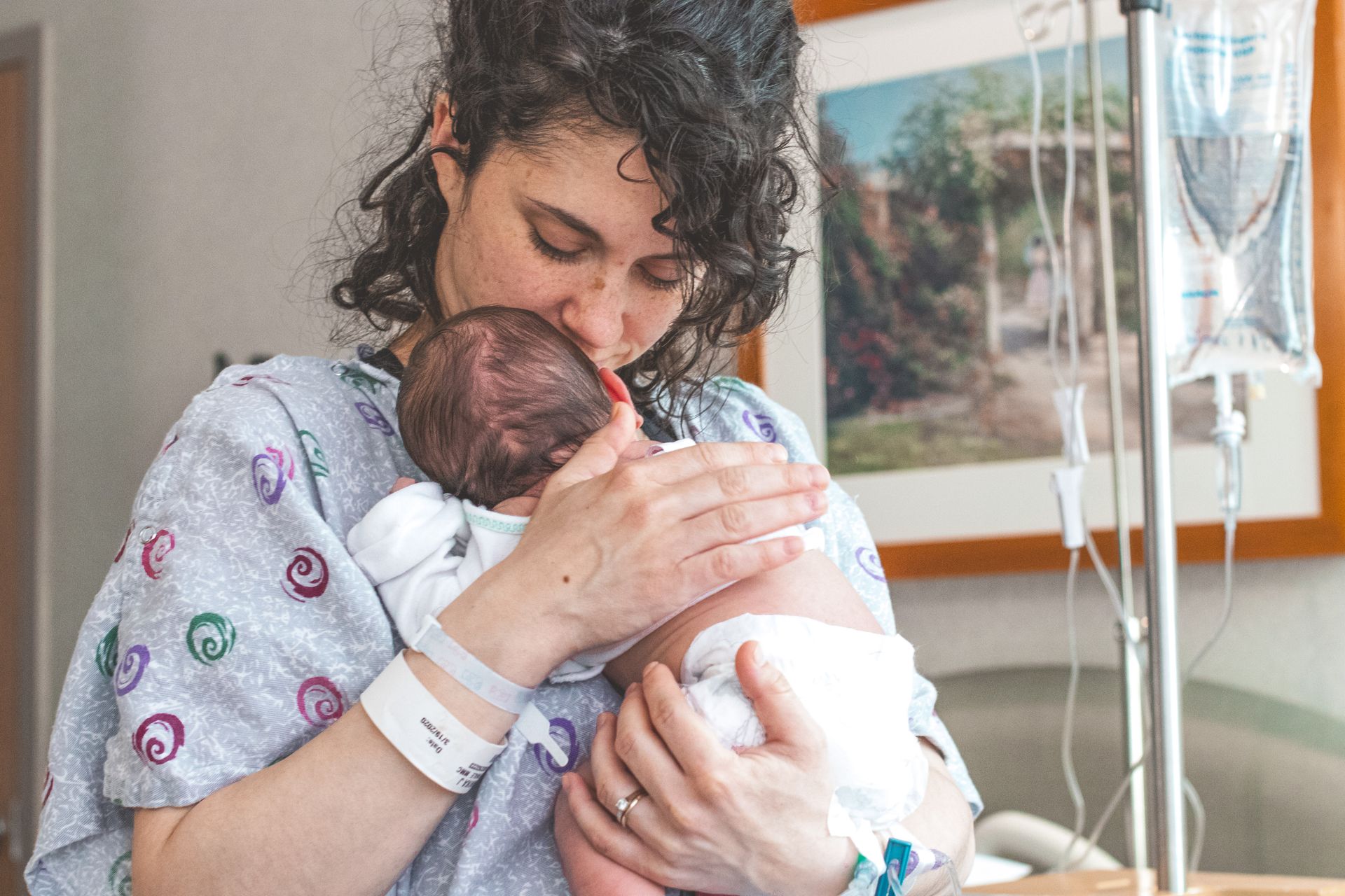 Mother Comforting Her Newborn After Childbirth Mother in a hospital gown holding her newborn against her chest beside an IV stand.