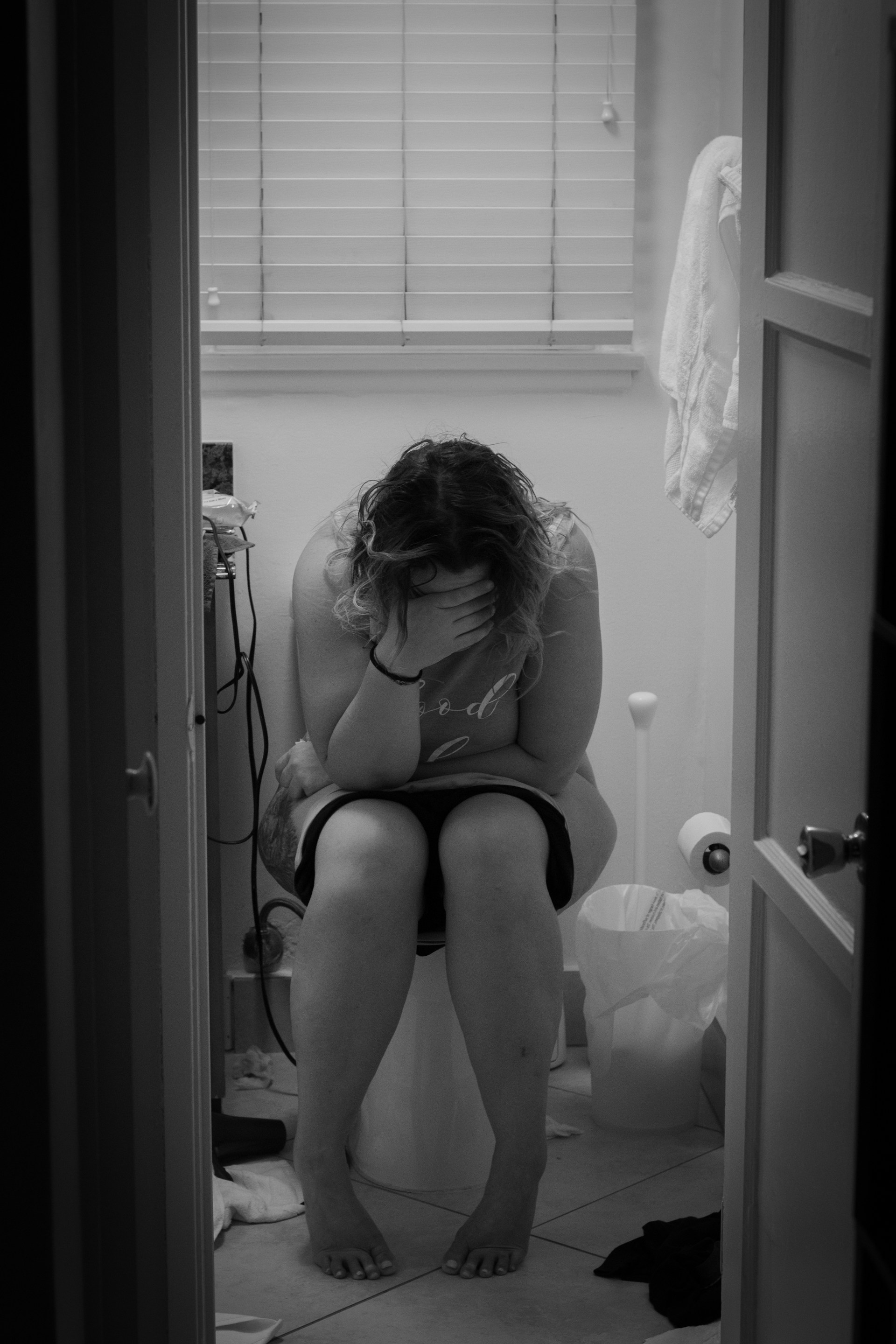 Physical Challenges During Postpartum Recovery Black-and-white photograph of a woman sitting in a bathroom and covering her face during postpartum recovery.