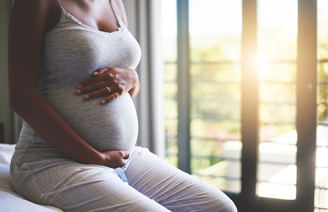 A pregnant woman sitting near a window, cradling her belly.