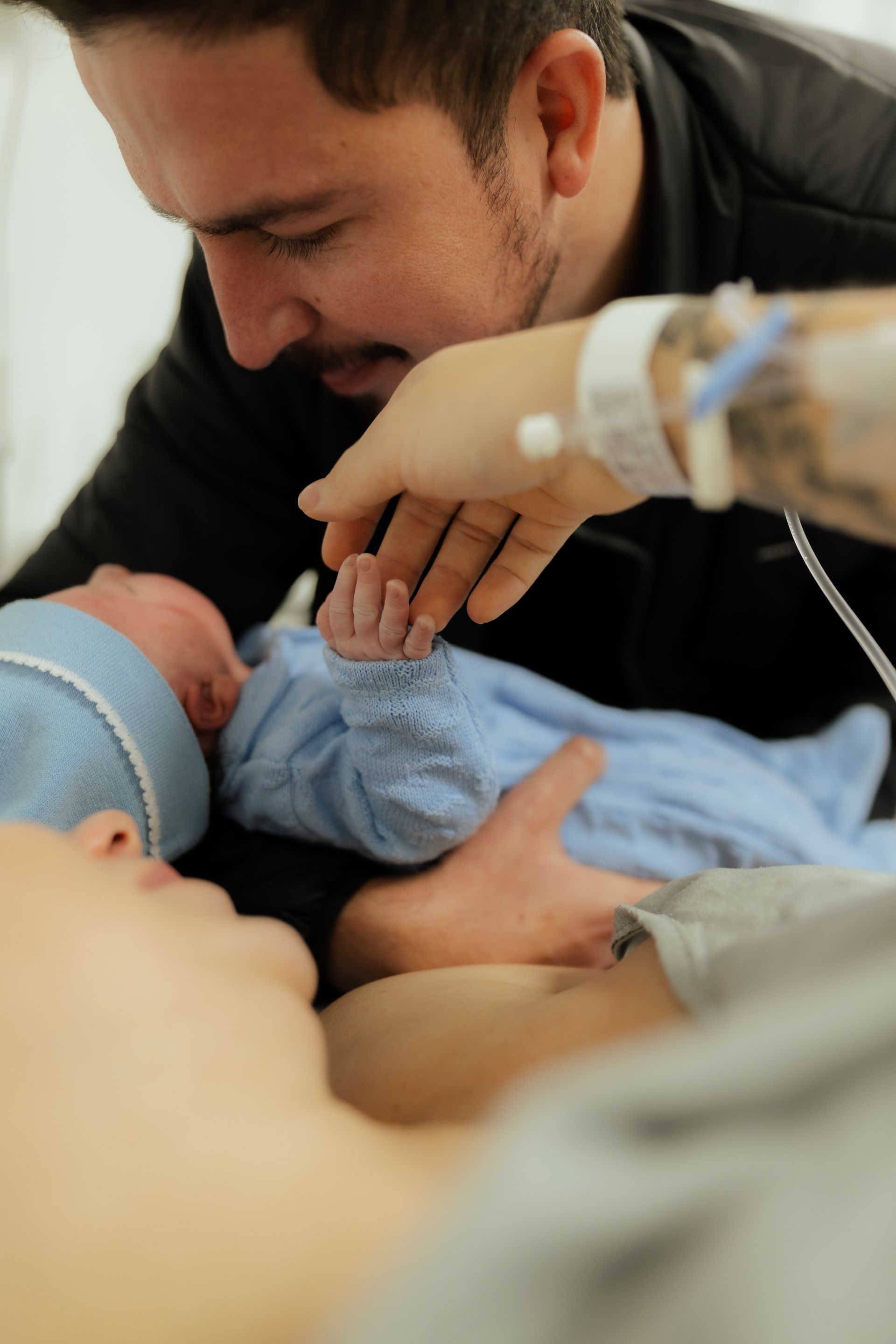 Parents Bonding With Their Newborn After Birth Father leans toward a newborn while the baby holds the hand of a parent receiving hospital care.
