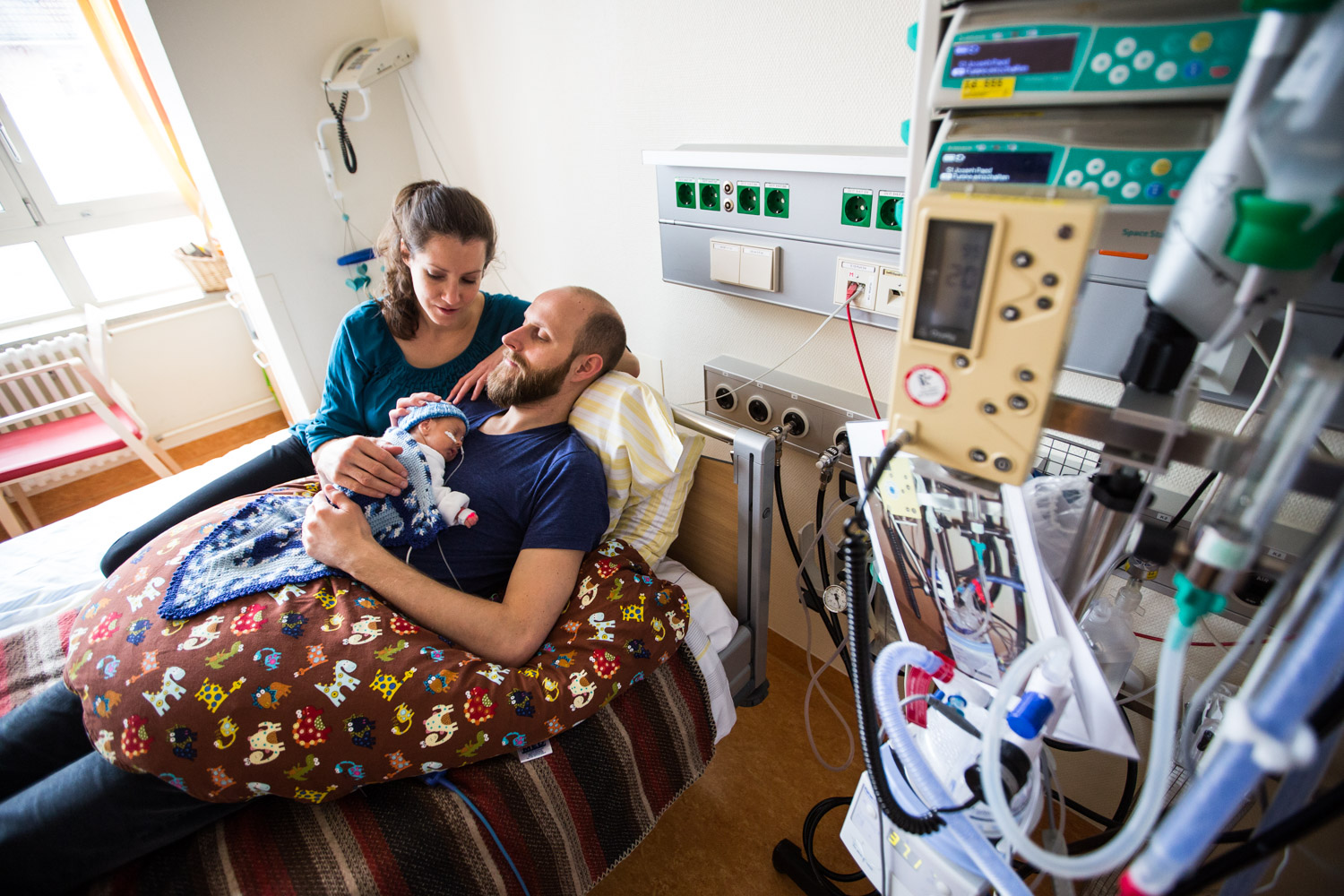 Parents Sharing Close Contact With Their Preterm Infant Mother and father sit together in a neonatal unit while the father holds their preterm infant against his chest.