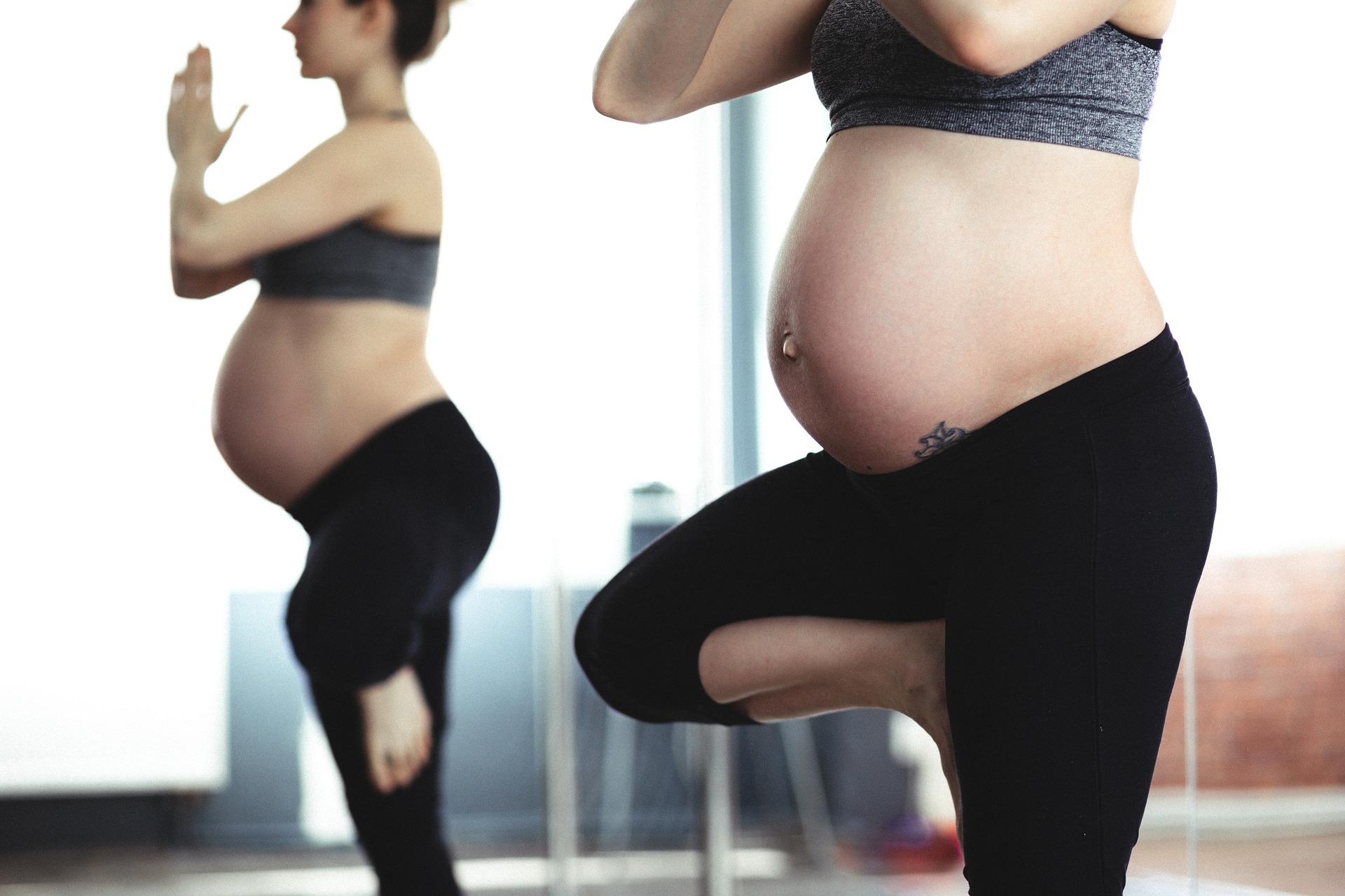 Prenatal Exercise and Maternal Wellbeing Two pregnant women wearing sportswear practice a standing yoga pose in a bright studio.