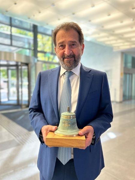 A man in a blue suit and tie stands indoors, smiling and holding a small bell trophy mounted on a wooden base, suggesting an award or recognition ceremony.