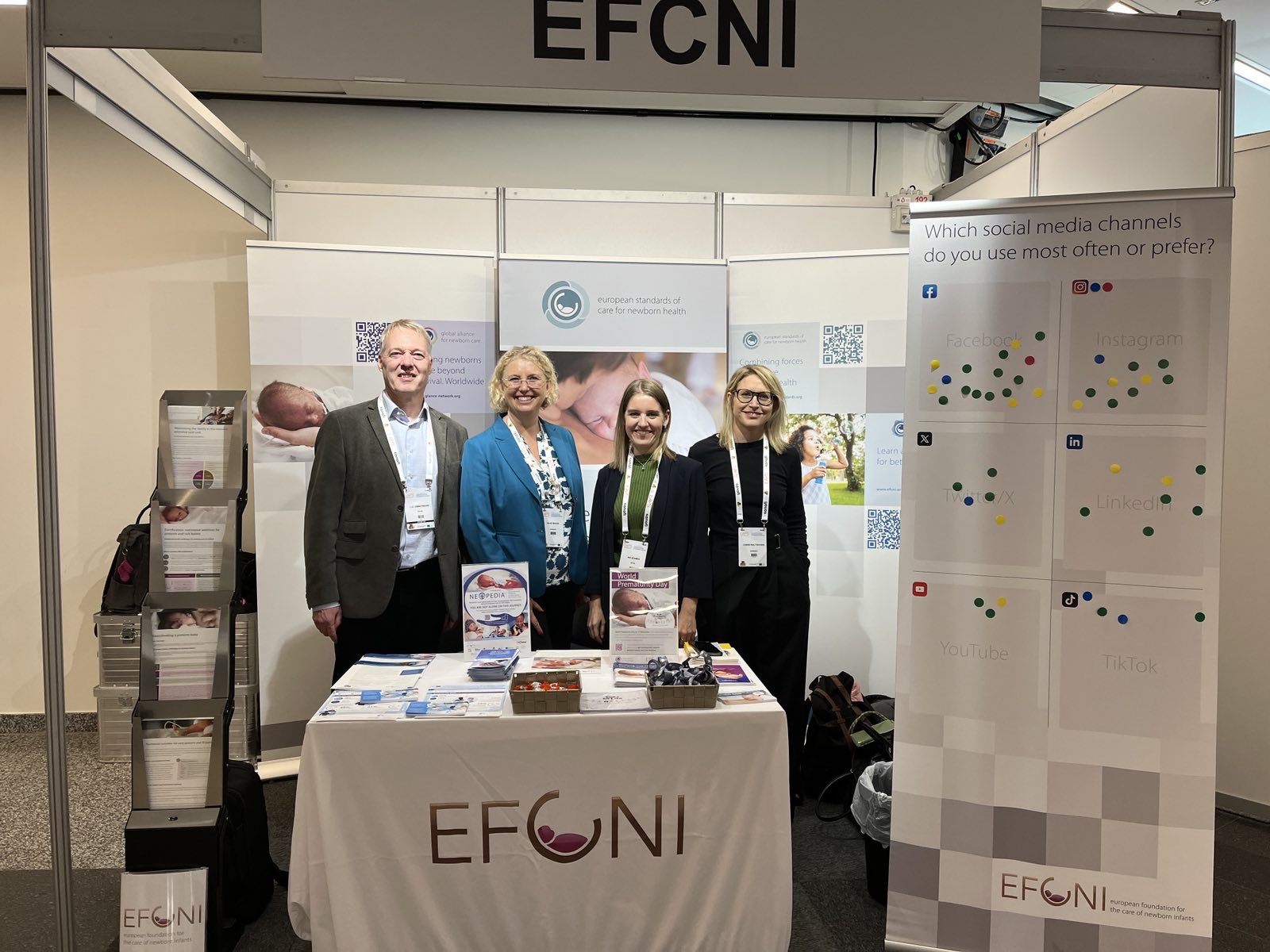 Group of four EFCNI team members standing at their booth, surrounded by informational material and a feedback wall on social media preferences.