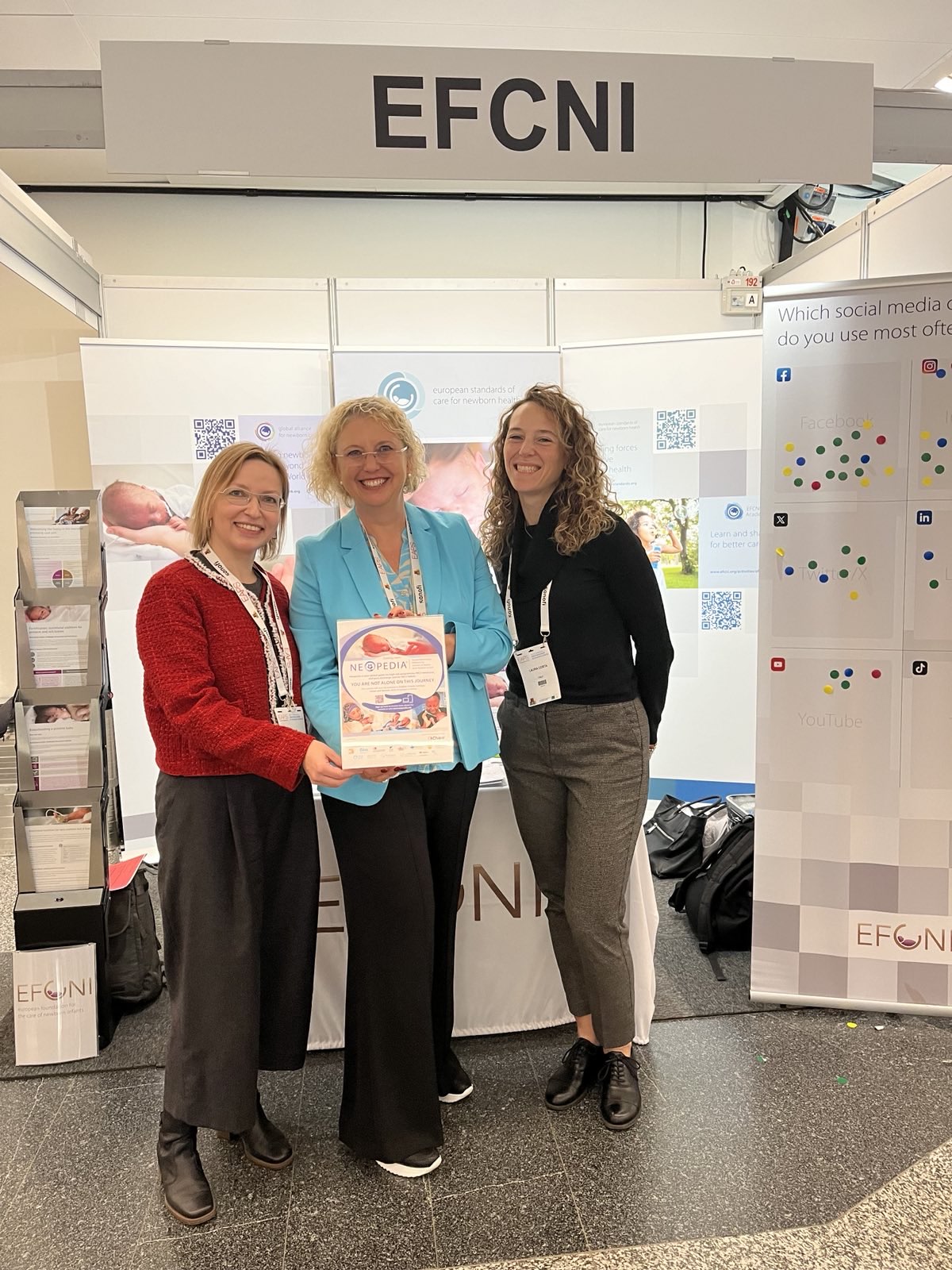 Three women from EFCNI at their exhibition booth, proudly presenting a NEO-PEDIA booklet focused on neonatal health and education.