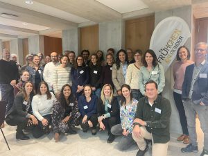 A large group of professionals pose together for a photo at a neonatal-focused event, standing in front of a “Frühchen” banner inside a modern conference space.