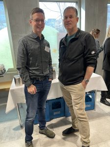 Two men wearing name tags stand by a table during a break at a neonatal care conference, casually chatting and smiling at the camera.