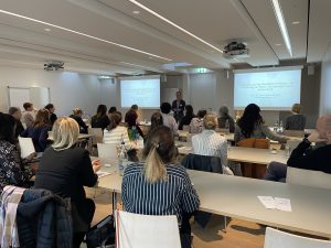 A room full of attendees listens to a speaker during a presentation session at a neonatal care event. Slides are projected on two screens.