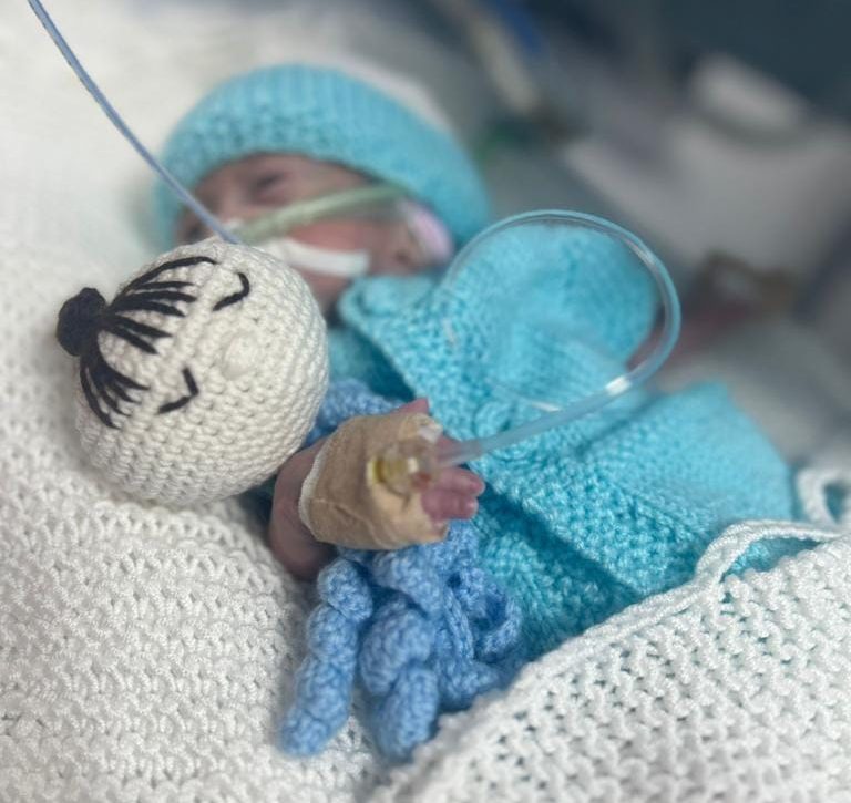 A premature baby lies in an incubator, dressed in a knitted blue outfit and holding a small crochet comfort toy, with oxygen tubes and a bandaged hand indicating intensive neonatal care.