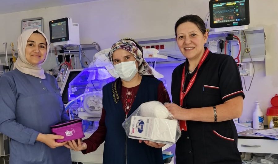 Three women stand smiling in a neonatal intensive care unit (NICU), with one holding a care package and another wearing a mask, surrounded by medical equipment and an incubator in the background.