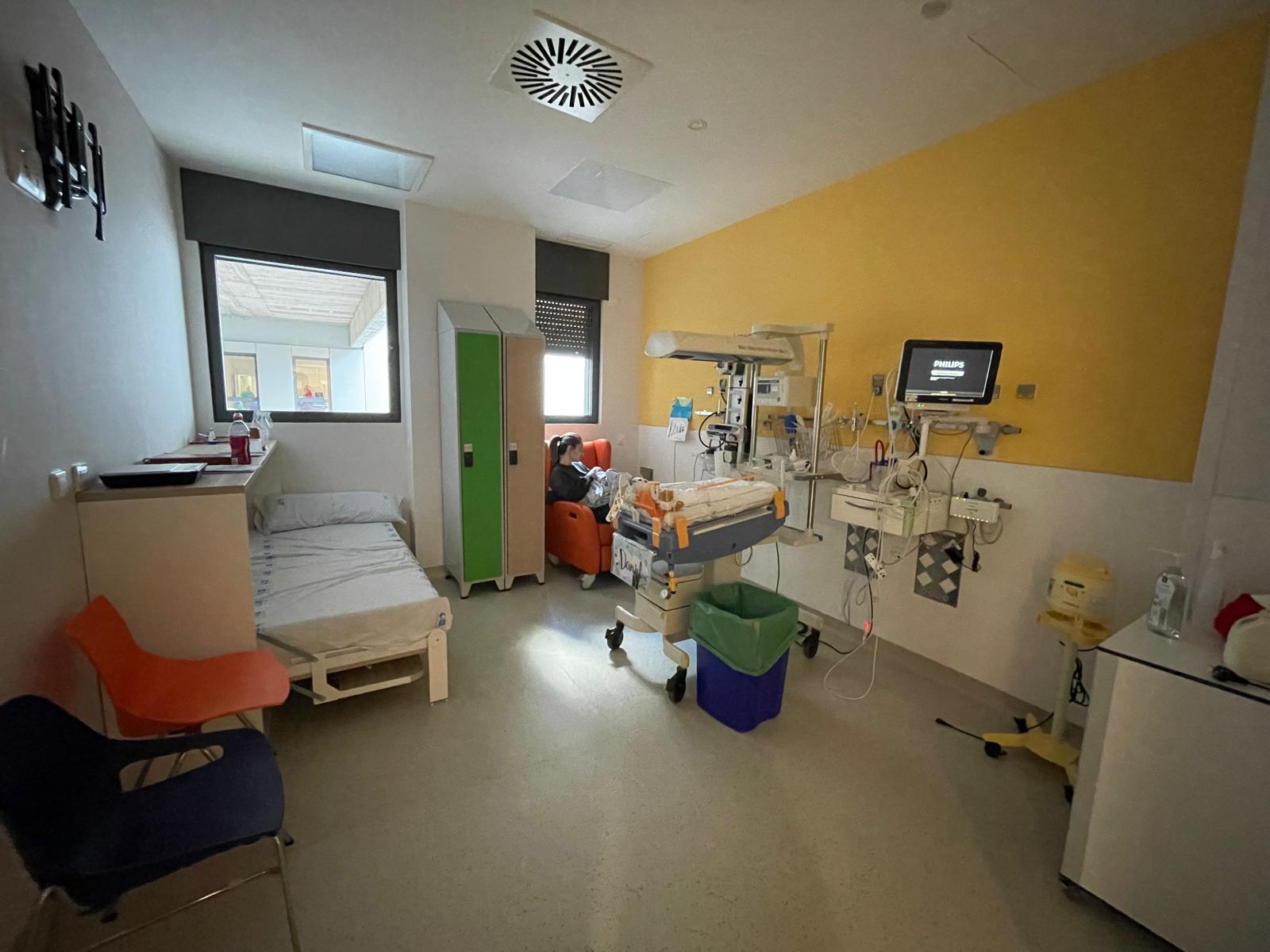 A modern hospital room equipped for neonatal care, featuring a neonatal intensive care unit (NICU) incubator, monitoring equipment, seating for parents, a hospital bed, and colorful furnishings creating a warm, family-friendly environment.