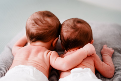 Two newborn twins lying close together on a soft surface, one gently resting an arm around the other, symbolizing closeness and early bonding.