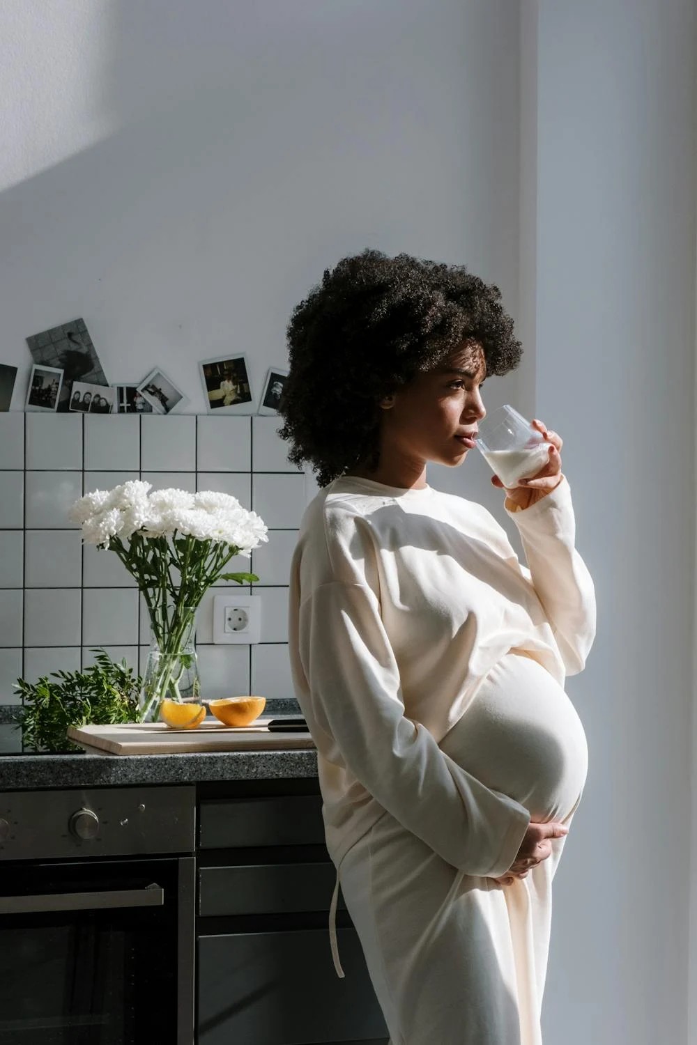 A pregnant woman in a cozy white outfit stands in a sunlit kitchen, gently holding her belly with one hand and sipping from a glass, with flowers and fruit on the counter behind her.